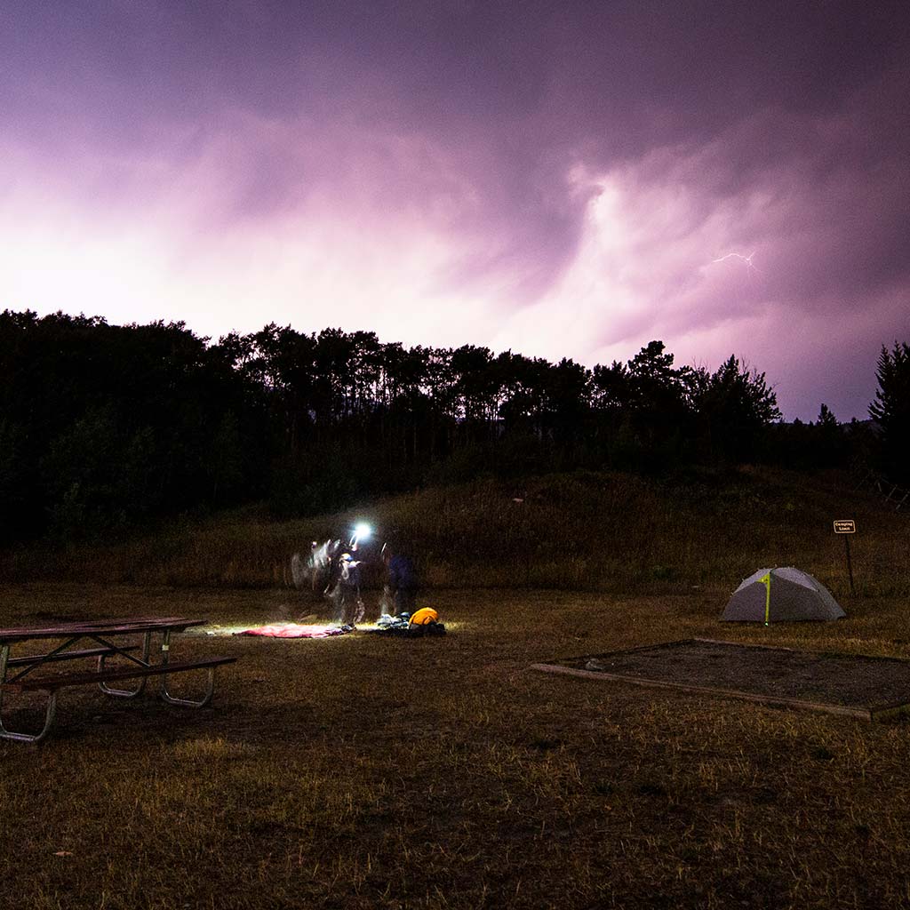 Lighting strikes, turning the sky white purple and pink. A tent and a picnic table are illuminated in the wide open space, trees line the space in the distance.