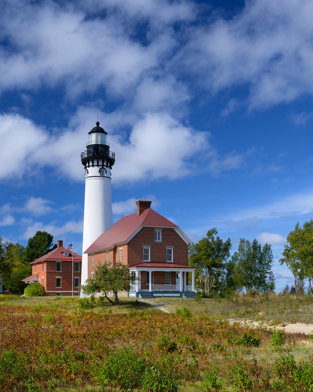 A white and brick lighthouse in a field of shrubs under a blue sky with white puffy clouds.