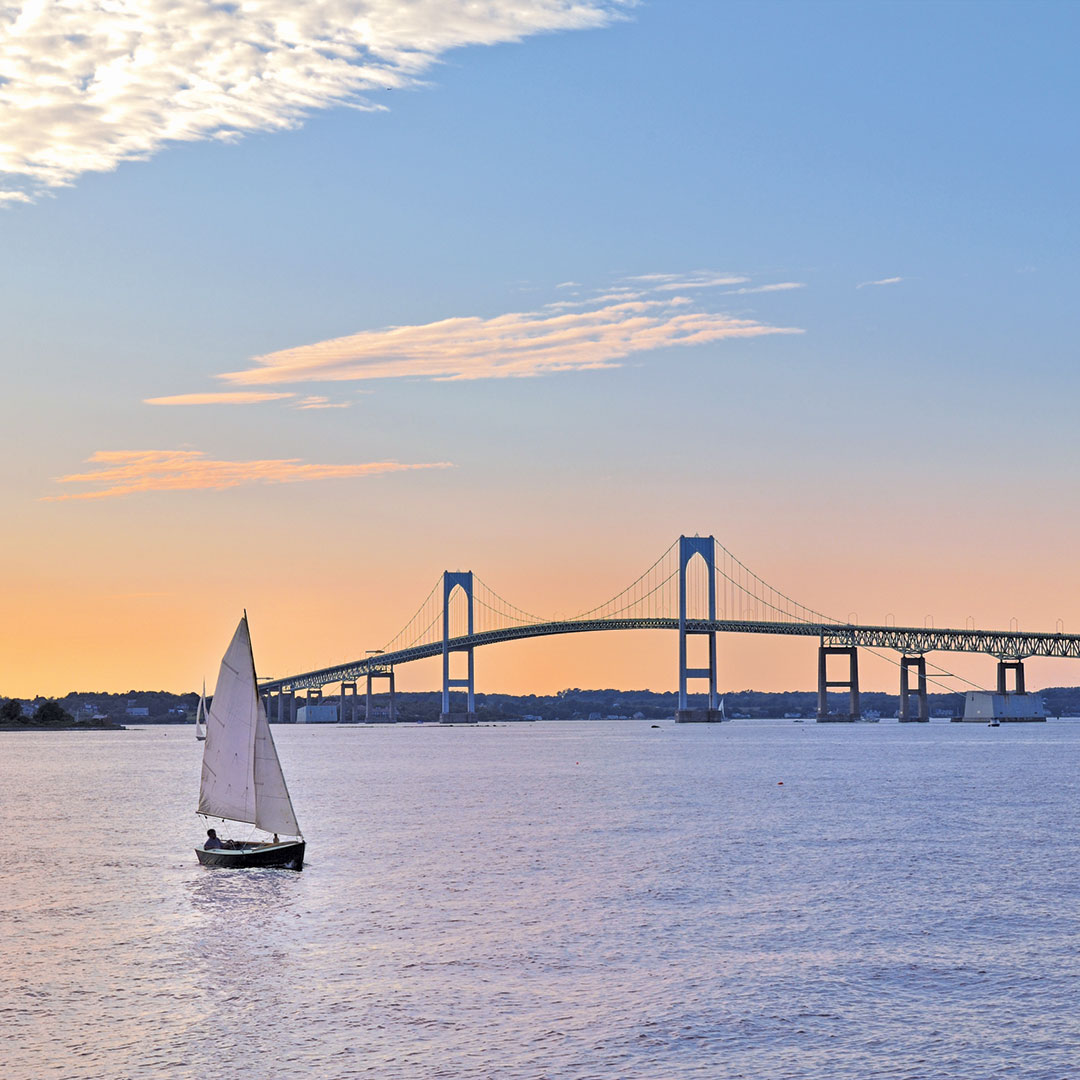 A sailboat in the water near Newport Bridge at twilight.